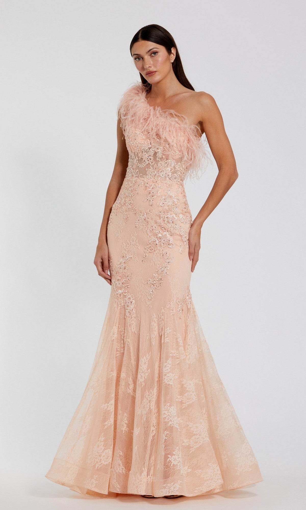 A woman models the Mac Duggal 79451 long formal dress in peach, featuring a mermaid silhouette, lace details, and a feathered one-shoulder neckline, standing against a plain white background.